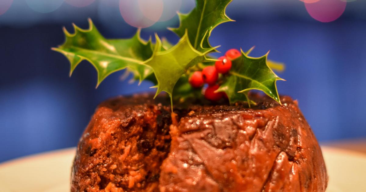 Christmas pudding with wreath on white plate and blue background, one slice taken out of it