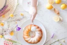 lemon bundt cake on white marble with white persons hand holding stainless steel sieve with icing sugar, lemons and flowers next to the cake too